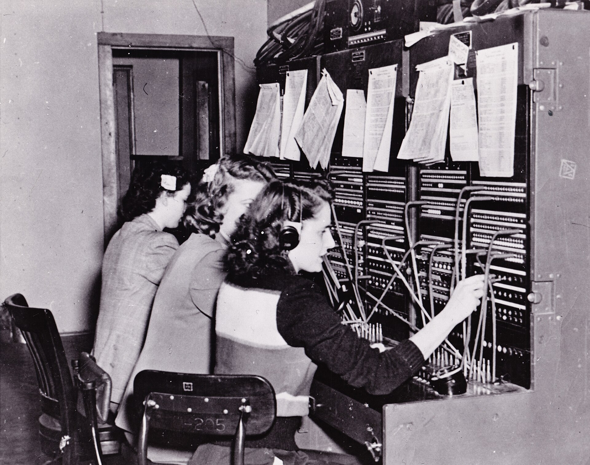 Three telephone operators at Fort Richardson's first switchboard in 1950, call records clipped in sheaves above the board.